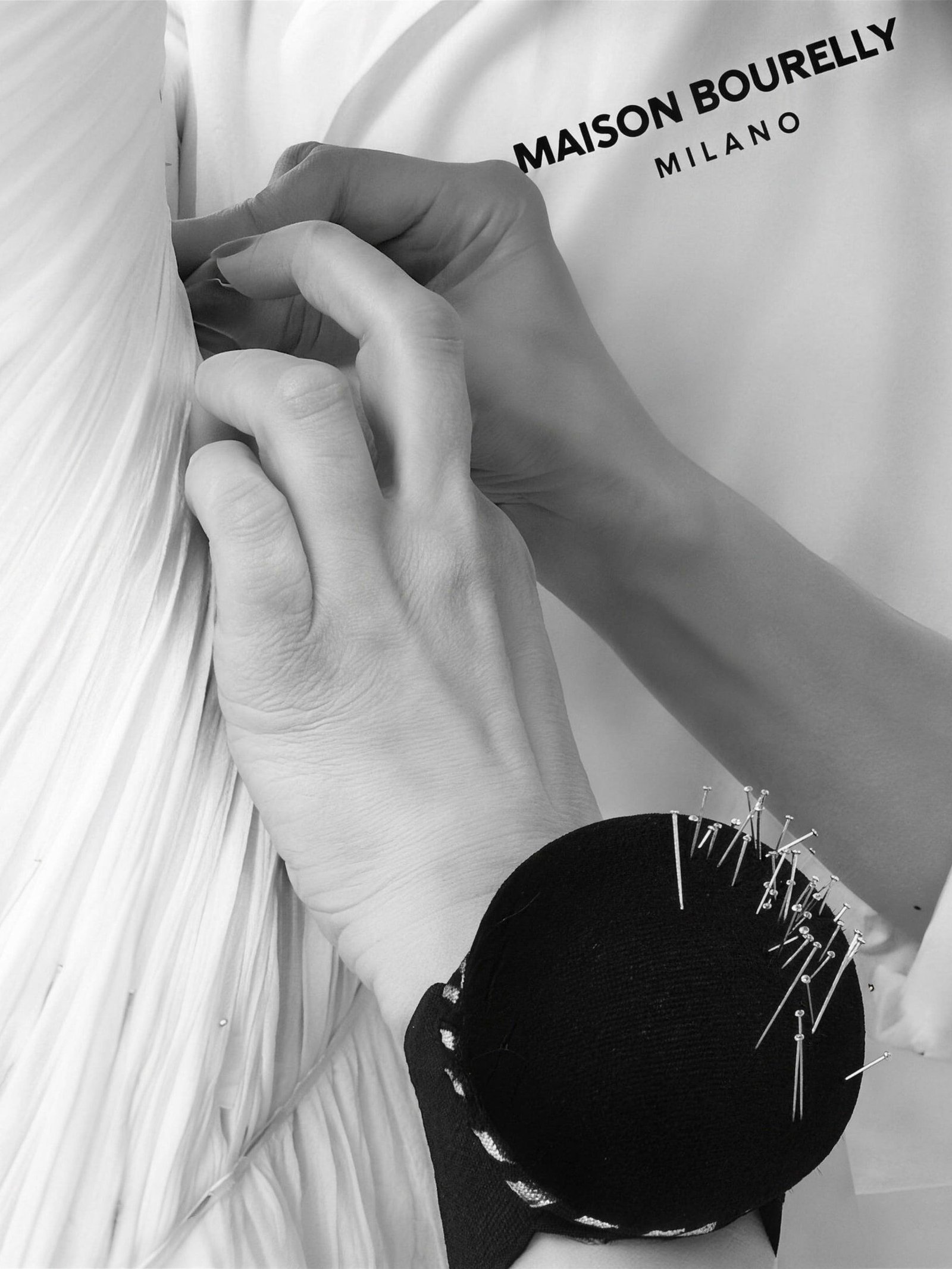 Mani che appuntano con gli spilli il plissé di un abito bianco, in atelier